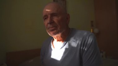 Evde yaşlı bir adam güneş ışığında yatak odasında yatağında oturuyor. Yalnızlık, yalnızlık, emeklilik kavramı. Yüksek kalite 4k görüntü