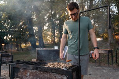 Orta yaşlı beyaz bir adam ızgara şampiyonları pişirirken kömürlere hava üflüyor. Açık havada mangal partisi, basit, lezzetli bir yemek. Yüksek kalite 4k görüntü