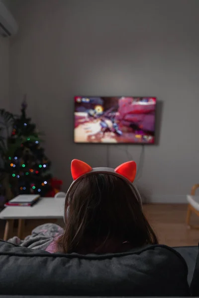 Back view of a child girl sitting on the couch and playing video game or watching cartoons on tv. Wireless funny pink headphones with ears. High quality photo