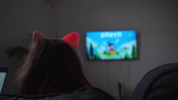 一个小女孩坐在沙发上玩电子游戏或看电视上的卡通片的背景图 带耳朵的无线有趣的粉色耳机 高质量的照片 — 图库视频影像