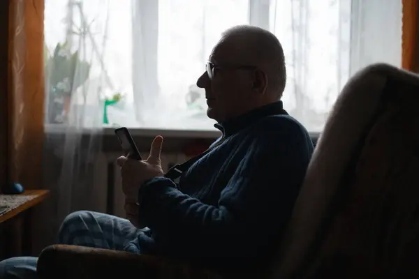 Senior male in arm sling looks at phone beside window
