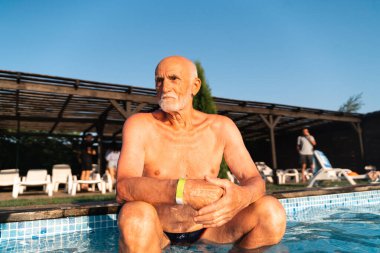 80 yaşında, sakallı, güneşli bir yaz gününde havuz kenarında dinlenen beyaz bir adam. Samimi yaşam tarzı, mutlu gerçek duygular. Yüksek kalite fotoğraf