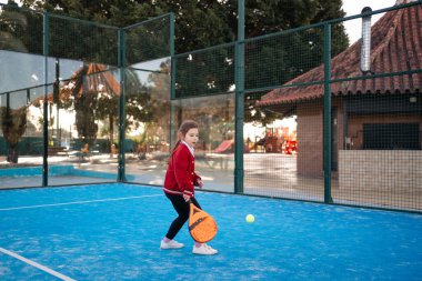 Odaklanmış genç bir kız açık bir günde canlı bir mavi kortta raket tenisi becerilerini geliştirir. Yüksek kalite fotoğraf