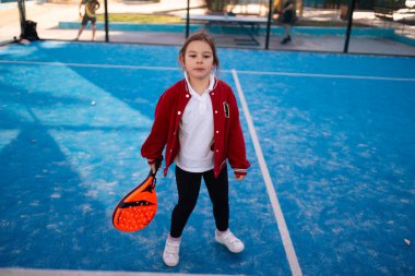 Kırmızı ceketli genç bir çocuk canlı turuncu bir padel raketin arkasına saklanıyor, kort ve arka planda bulanık oyuncular var. Yüksek kalite fotoğraf