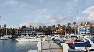 Sotorande İspanya - 25 Mart 2024 Renkli evler ve palmiye ağaçlarıyla kaplı gösterişli bir marina, yumuşak mavi gökyüzü altında sofistike yatlar ve tekneler sergiliyor. Yüksek kalite 4k görüntü