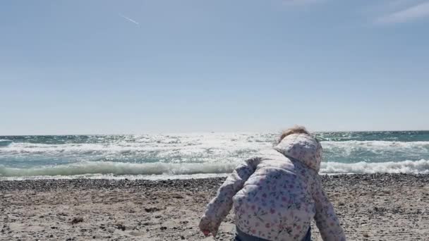 一个年轻的孩子 背对着摄像机 正在探索一个被卵石覆盖的海滩 背景中的波浪轻轻破碎 高质量的4K镜头 — 图库视频影像