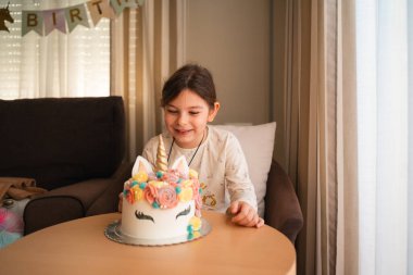 Neşeli bir doğum günü kızı, hayallerindeki tek boynuzlu at kekine dokunmak için uzanırken gülümsüyor. Kutlamasının unutulmaz bir anı. Yüksek kalite fotoğraf