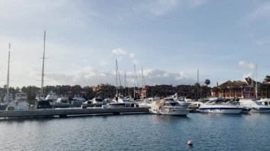 Sotorande İspanya - 25 Mart 2024 Renkli evler ve palmiye ağaçlarıyla kaplı gösterişli bir marina, yumuşak mavi gökyüzü altında sofistike yatlar ve tekneler sergiliyor. Yüksek kalite 4k görüntü