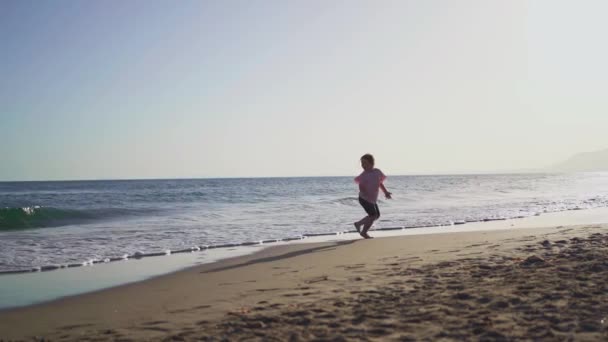 一个年轻姑娘高兴地沿着海滨跑着 日落时海浪拍打着海岸 高质量的4K镜头 — 图库视频影像