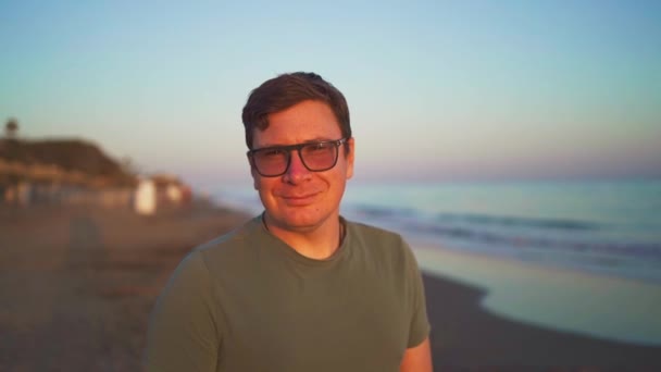 一个快乐的男人 在夕阳西下 带着眼镜在模糊的海滩背景下微笑 散发出幸福和放松的光芒 高质量的4K镜头 — 图库视频影像