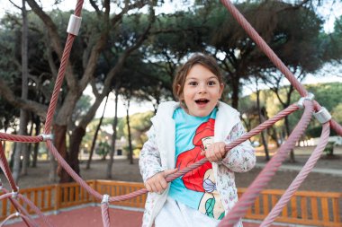 Coşkulu genç bir kız parktaki bir halat yapısına tırmanmayı seviyor, mutluluk ve gençlik enerjisi yayıyor. Yüksek kalite fotoğraf