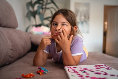 Genç bir kız, oyuncaklarla çevrili bir kanepede yatarken esprili bir şekilde komik suratlar yapar. Yüksek kalite fotoğraf
