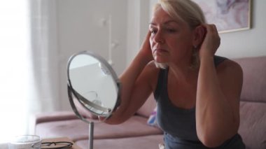  Son sınıf öğrencisi bir kadın, oturma odasında otururken aynada saçına dikkatlice bakıyor, özbakım ve yaşlanmayı yansıtıyor. Yüksek kalite 4k görüntü