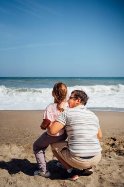 Güneşli bir kumsalda dururken gülücüklerini ve kahkahalarını paylaşan bir baba ve kız arasında neşeli bir an. Yüksek kalite fotoğraf