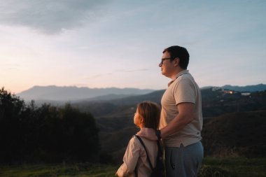 Bir baba, akşam gökyüzünün altındaki tepelerin ve dağların manzarasının tadını çıkarırken kızını kucaklıyor. Yüksek kalite fotoğraf
