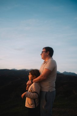 Bir baba, akşam gökyüzünün altındaki tepelerin ve dağların manzarasının tadını çıkarırken kızını kucaklıyor. Yüksek kalite fotoğraf