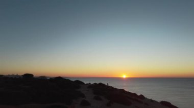  Siluetli figür dramatik uçurumlarda canlı bir Algarve gün batımı yakalıyor. Solo seyahat ve manzara fotoğrafçılığı için ideal sakin bir an. Yüksek kalite 4k görüntü