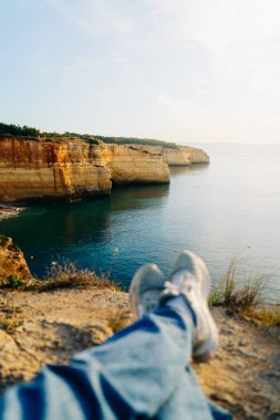 Portekiz 'in Algarve kayalıklarında rahatlayan birinin kişisel perspektifi. Altın kireçtaşı kayalıkları parlak, güneşli bir gökyüzünün altında sakin mavi sularla buluşur. Yüksek kalite fotoğraf