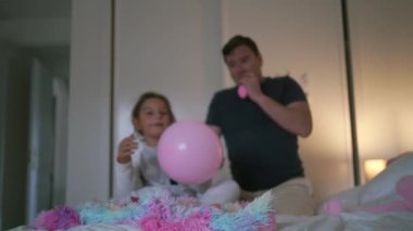 Bir baba ve kızı, renkli battaniyeler ve tek boynuzlu at dekoru ile çevrili bir yatakta parlak bir yatak odasında balonlarla şakalaşarak etkileşim içinde. Yüksek kalite 4k görüntü