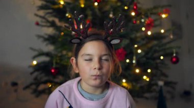 Geyik boynuzu takan gülümseyen bir çocuk süslerle süslenmiş parıldayan bir Noel ağacının önünde poz veriyor. Yüksek kalite 4k görüntü