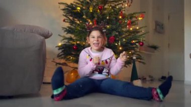 Şen şakrak giyinmiş bir çocuk süslü bir Noel ağacının önünde yere oturup heyecanını gösteriyor. Yüksek kalite 4k görüntü