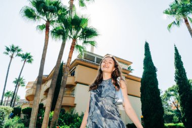 Akan gri elbiseli genç bir kadın, arka planda modern bir binayla palmiye ağaçları arasında yürürken parlak bir şekilde gülümsüyor. Yüksek kalite fotoğraf