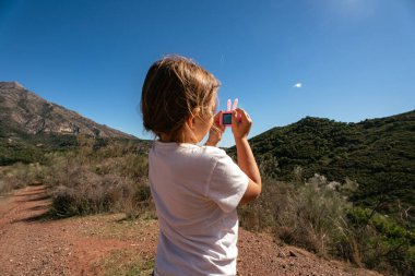 Young caucasian child taking photos in mountain landscape on a clear day. clipart