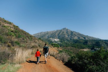 Baba ve oğul açık mavi gökyüzünün altında dağ yolunda yürüyüş yapıyorlar..