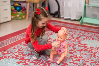 Kırmızılı küçük bir kız bir bebekle oynanıyor. Oyuncak bebeği evde halının üzerine koyuyor.