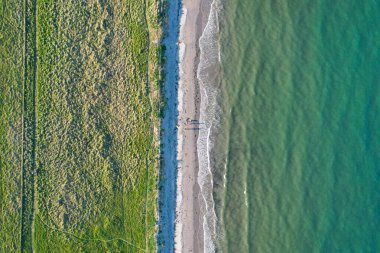 İrlanda 'da yeşil kum tepecikleri ve turkuaz deniz ile buluşan kumlu bir sahilin yukarıdan aşağı hava görüntüsü. İnsanlar akşam ışığında uzun gölgeler bırakıyorlar..