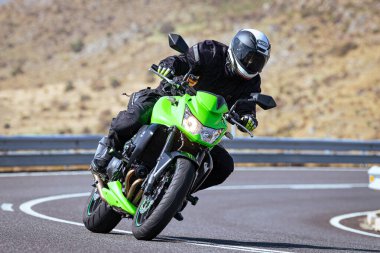 Motosiklet sürücüsü, arka planı doğa ve mavi gökyüzü olan ön planda bir viraj alıyor.