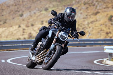 Motosiklet sürücüsü, arka planı doğa ve mavi gökyüzü olan ön planda bir viraj alıyor.