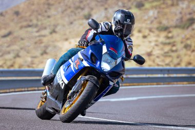 Motosiklet sürücüsü, arka planı doğa ve mavi gökyüzü olan ön planda bir viraj alıyor.