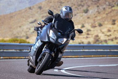 Motosiklet sürücüsü, arka planı doğa ve mavi gökyüzü olan ön planda bir viraj alıyor.