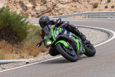 Yoldaki virajda motosikletini süren genç bir adam, ön planda güneşli bir günde fotoğraf. Fotoğraf Ağustos 2021 'de İspanya' nın Avila ilinde çekildi.