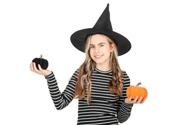 Cadılar Bayramı konsepti. Siyah şapkalı gülümseyen bir kızın portresi beyaz arka planda dekoratif balkabakları tutuyor. Cadılar Bayramı Dekorasyonu.