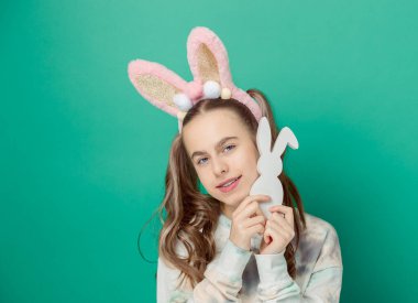 Mutlu Paskalyalar. Mutlu gülüşü olan, parlak pembe tavşan kulakları takan ve elinde ahşap bir tavşancık figürü olan, mavi arka planda Paskalya eğlencesi saçan bir kız. Dişlerinde diş telleri olan sevimli bir gülümseme..