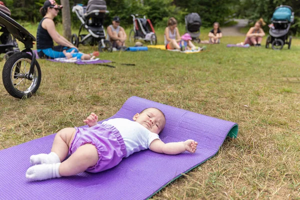 Wide angle shot of newborn baby laying on a purple yoga mat, on the grass with its parents sitting with their infants in the background