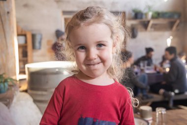 Kıvırcık sarı saçlı, kırmızı gömlekli, meşgul bir kafede duran gülümseyen genç bir çocuğun portresi. Seçici odak