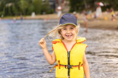 Uzun saçlı genç bir çocuk güneşli bir yaz gününde gölde oynuyor, can yeleği giyiyor ve gülümsüyor. Seçici odak