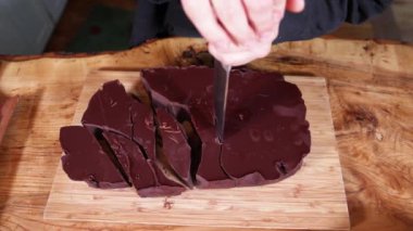 Çikolatacı dikkatle bir parça siyah çikolatayı tahta kesme tahtasında küçük parçalara ayırıyor. Seçici odak