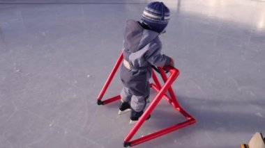 Buz pateni öğrenen küçük bir çocuk, dışarıda, karlı bir buz pateni pistinde kırmızı bir yürüteçle destek veriyor. Seçici odak