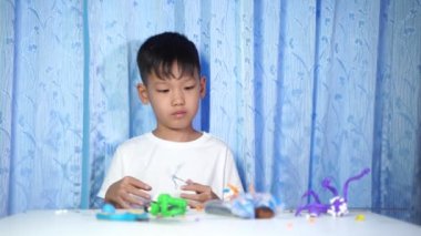 Konsantre olmuş Asyalı çocuk evdeki beyaz bir masada oturup plastik bebeklerden yapıyor.