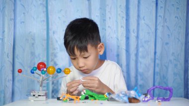 Konsantre olmuş Asyalı çocuk evdeki beyaz bir masada oturup plastik bebeklerden yapıyor.