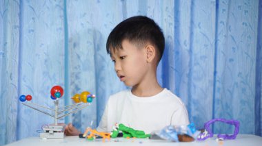 Konsantre olmuş Asyalı çocuk evdeki beyaz bir masada oturup plastik bebeklerden yapıyor.