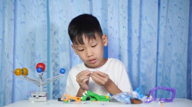 Konsantre olmuş Asyalı çocuk evdeki beyaz bir masada oturup plastik bebeklerden yapıyor.