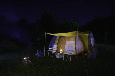 Geceleri yeşil çimlerin üzerinde kamp çadırı, antika gaz lambaları, retro sandalyeler, ormandaki kamp alanındaki masalarda açık kahve yapma tesisleri.