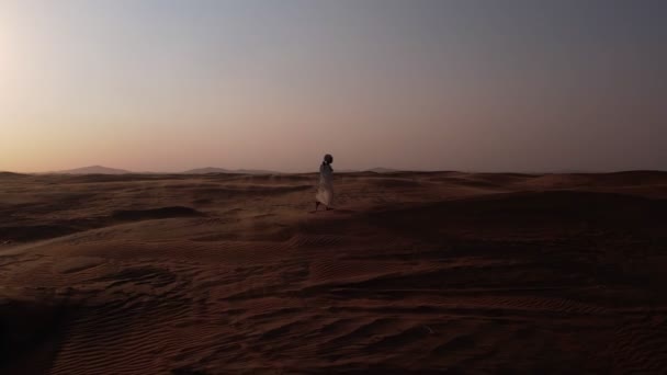 无人驾驶飞机发射了一个身穿飘扬的白色长袍和头巾的瘦弱的阿拉伯男子 光着脚穿过沙漠走向日落 — 图库视频影像
