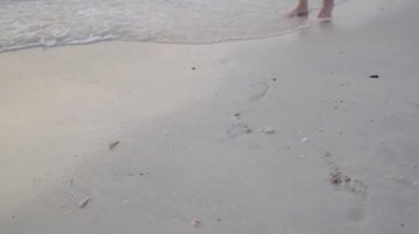 Gün batımında denizin kumlu kıyılarında yürüyen açık renk elbiseli genç bir kadının ayakları.