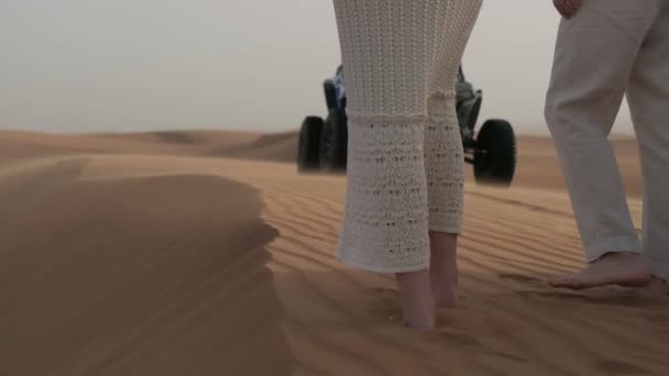 男と女はバギーを背景に手を取りながら砂漠を裸足で歩く — ストック動画